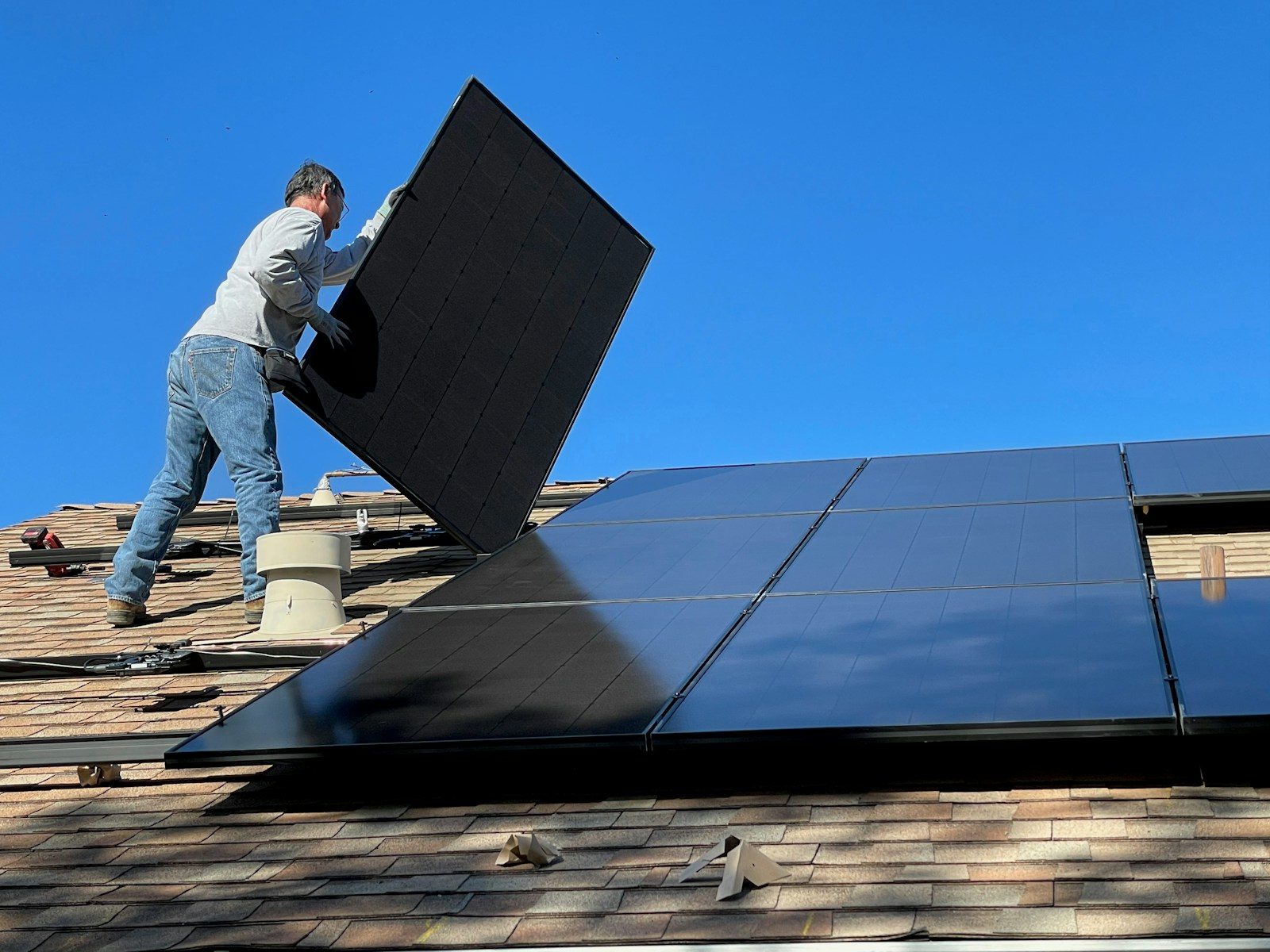 A person installing a black solar panel on a shingled roof under blue sky